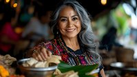 Mature woman with long gray hair and South Asian ethnicity wearing traditional patterned clothing, preparing fresh vegetables in a home kitchen setting with warm lighting.