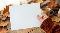 Caucasian hand in rust-colored knit sleeve holding blank white card over wooden surface with autumn leaves for greeting, invitation, or seasonal marketing mockup.