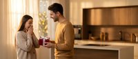 Young Caucasian man presenting gift box to surprised woman in modern kitchen during romantic moment at home.