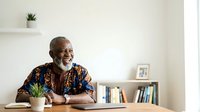 Senior African American man with gray beard wearing colorful patterned shirt, sitting at desk with potted plant and bookshelf, smiling while working in bright modern office space.