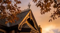 Victorian house gable with ornate wooden trim and decorative finial against warm sunset sky framed by autumn tree branches creating architectural detail focus.
