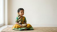 Young South Asian girl in traditional green and gold silk attire with white flower garland, seated on woven mat by window, cultural celebration or festival portrait.