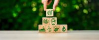 Hand placing wooden cube with target symbol on top of eco-friendly icons pyramid including recycling, renewable energy, and nature symbols on green background.