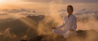 Young Asian man in white clothing meditating on mountain peak at sunrise with golden sunbeams piercing through misty clouds and dramatic landscape below.