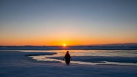 Solitary figure walking on frozen lake at sunset with golden sky reflection on ice surface creating peaceful winter landscape scene for meditation concepts.