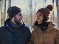Cheerful interracial couple in winter clothing sharing genuine laughter outdoors during cold season, wearing knit beanies and warm jackets against blurred forest background.