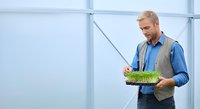 Middle-aged Caucasian male agronomist or farmer examining seedling tray with green sprouts in greenhouse environment, wearing blue shirt and vest for agricultural research and cultivation.
