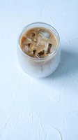 Iced coffee with cream swirl in glass mug on white wooden surface, refreshing cold beverage for summer morning cafe menu and restaurant advertising.