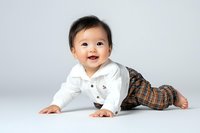 Adorable Asian baby boy crawling on white background, wearing white shirt and plaid pants, smiling joyfully. Studio portrait for parenting content.