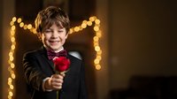 Young boy in formal black tuxedo holding red rose with warm bokeh lights creating romantic heart shape background for special occasions and celebrations.