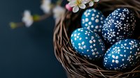 Decorative blue Easter eggs with white floral pattern in woven nest basket, with delicate spring blossoms against dark background. Traditional holiday decor.