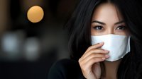 Young Asian woman wearing protective face mask looking at camera with intense eyes against blurred background with warm bokeh light effect.