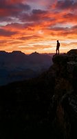 Silhouetted male hiker standing on rocky cliff edge overlooking vast canyon landscape during dramatic golden and orange sunset. Inspiring adventure and exploration concept.