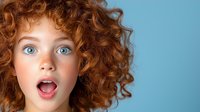Young girl with bright blue eyes, natural red curly hair and freckles expressing surprise and wonder against light blue background. Close up portrait showing genuine emotion.