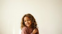 Young girl with curly blonde hair and light skin tone wearing a dusty rose colored shirt, smiling at camera against neutral background for portrait and family use.