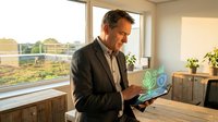 Middle-aged man with dark hair and light skin tone, wearing business casual attire, holding laptop displaying green sustainability icons in modern office with natural light and plants.