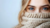Young woman with striking blue eyes and blonde hair partially covered by cozy cream knit turtleneck sweater against light background. Close up portrait showing natural beauty.