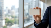 Male professional in business attire holding blank white card against urban cityscape window background, suitable for corporate branding, business card mockups, and professional presentations.