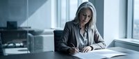 Professional businesswoman with gray hair working at desk in modern office, reviewing documents and paperwork for corporate meeting preparation.