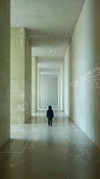 Person in dark clothing standing alone in minimalist corridor with white walls and geometric floor patterns, representing solitude, isolation, or contemplation in modern architecture setting.