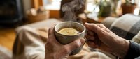 Senior man with weathered hands holding warm herbal tea in ceramic bowl, relaxing at home with cozy blanket and plants in background for wellness and lifestyle content.