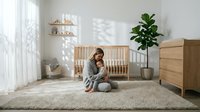 Young Caucasian woman in casual gray clothing sitting peacefully on soft rug in modern minimalist nursery with wooden crib and natural decor elements.