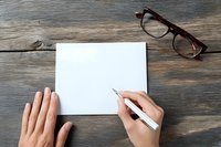 Hands writing on blank white paper with pen on rustic wooden desk surface with eyeglasses nearby for office work and creative projects.