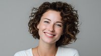 Happy young Caucasian woman with curly brown hair smiling warmly at camera wearing white top against neutral gray background for professional headshot.