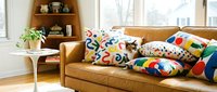 Tan leather sofa with colorful patterned pillows in bright living room with wooden shelving, white side table, potted plant, and natural window light for home decor.