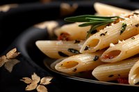 Cooked penne pasta with herbs and spices on black plate, garnished with fresh rosemary sprigs and golden leaf decorations, dramatic food photography.