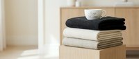 Stack of folded soft blankets in neutral tones with a white cup on top in a cozy minimalist interior setting.
