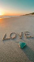 Word LOVE written in sand on a sunset beach with a turquoise sea glass heart, romantic coastal scene for valentines and travel.