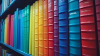 Colorful hardcover books arranged in rainbow order on wooden library shelf, spines creating vibrant gradient pattern. Educational and decorative background.