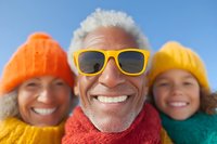 Cheerful mature man with gray beard wearing yellow sunglasses and red sweater taking selfie with family in colorful winter knit hats, showing genuine happiness and connection.