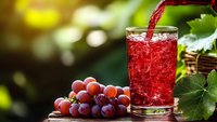 Fresh red grape juice being poured into glass with ice cubes and grape cluster on wooden table in sunny garden setting for healthy beverage concepts.