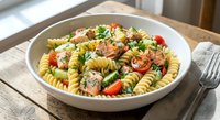 Delicious pasta salad with spiral noodles, cherry tomatoes, fresh herbs and vegetables served in white bowl on rustic wooden table for healthy meal preparation.