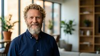 Mature Caucasian businessman with curly hair and beard smiling confidently in modern office interior with natural lighting and plants.