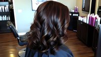 Luxurious dark brown hair with soft waves and highlights styled in loose curls showcases modern salon hairstyling technique on wooden floor background in beauty salon.