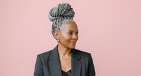 Mature African American woman with gray braided hair styled in an updo, wearing a black blazer over a black top, smiling confidently against a pink background. Corporate portrait for business, professional headshot, and leadership themes.