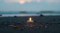 Single lit candle glowing on dark sandy beach at dusk, with blurred ocean waves and setting sun in background creating peaceful meditation atmosphere.