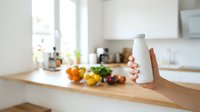 Hand holding white blank bottle with fresh vegetables and fruits on kitchen counter, promoting healthy eating, nutrition products, organic food supplements, wellness lifestyle and home cooking.