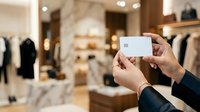Person holding blank white credit card in upscale clothing boutique with marble flooring and designer garments displayed on racks and shelves for retail shopping and payment concepts.