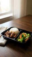 Healthy takeout meal in black container with glazed meat, steamed rice and fresh vegetables on wooden table near window for delivery service.