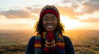 Young African woman wearing traditional colorful knit hat and sweater smiling at golden hour sunset with warm landscape background for cultural diversity content.