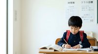 Young East Asian boy in school uniform writing in notebook at desk, educational setting with whiteboard background.