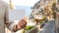 Caucasian male holding blank white card with coastal village and Mediterranean seascape in background, suitable for travel marketing, tourism promotions, and vacation advertising campaigns.
