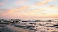 Soft ocean waves roll onto sandy shore during golden hour sunset, creating serene coastal landscape with warm pastel sky tones. Perfect for travel, nature, and relaxation themes.