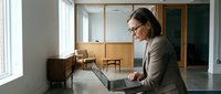 Professional businesswoman working on laptop in modern office space with natural lighting and minimalist wooden furniture for corporate productivity.