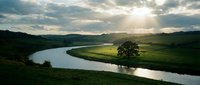 Scenic river landscape with lush green fields, rolling hills, and dramatic sky with sunlight breaking through clouds, suitable for nature, travel, and environmental design projects.
