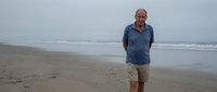 Mature man with glasses and beard wearing casual blue shirt standing alone on empty sandy beach with ocean waves in background during overcast day.
